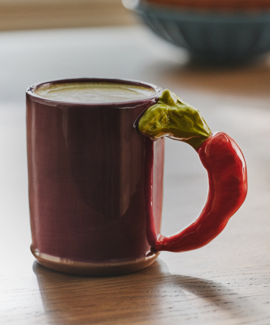 Brown ceramic mug with red chili pepper-shaped handle and green leaf accent, filled with coffee on wooden table