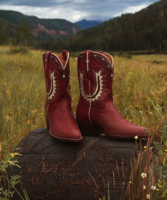 Red cowboy boots with white decorative stitching on wooden platform in golden field with mountains in background