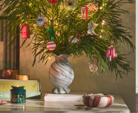 Decorated evergreen branches in a white marbled vase with colorful ornaments, candy canes, and lights on a white surface.