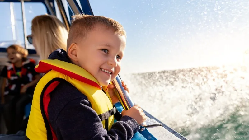Portrait of cite little blond happy excited smiling caucasian boy wear lifevest enjoy sailing on motor boat sea against blue sky and water splash wave sun backlit.