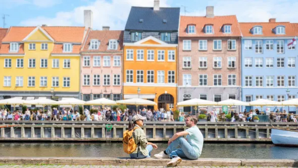 Ein Mann und eine Frau sitzen in Nyhavn in Kopenhagen auf der Hafenmauer mit bunten Häusern im Hintergrund.