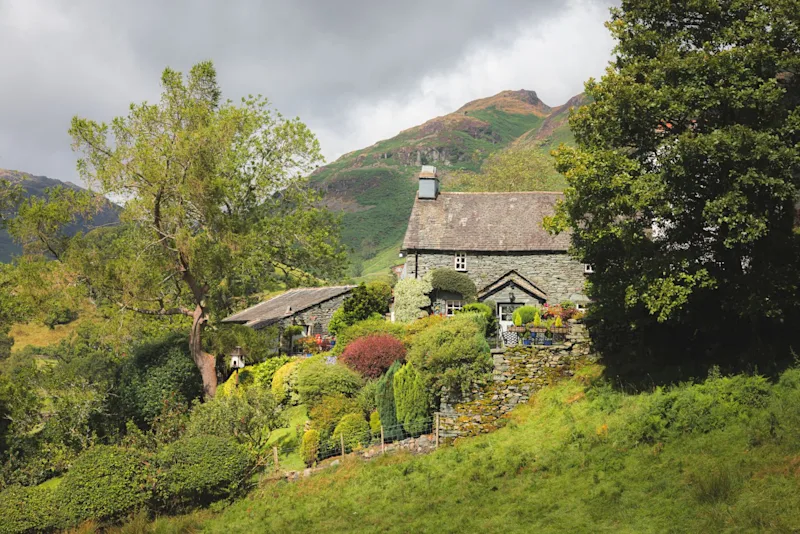 A quaint stone cottage in Langdale, near Ambleside, is surrounded by a serene garden and greenery, set against the picturesque Lake District mountains