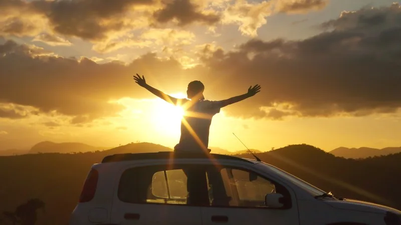 Happy and free man standing through a car sunroof at golden hour, arms open to the sky. Concept of freedom, gratitude, road trip, slow travel