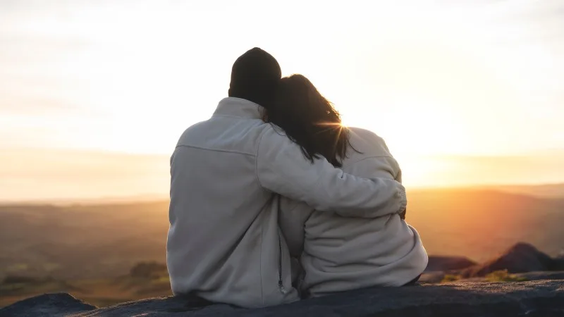 Back view of the happy couple in love sitting on top of a mountain enjoying a sunset landscape view