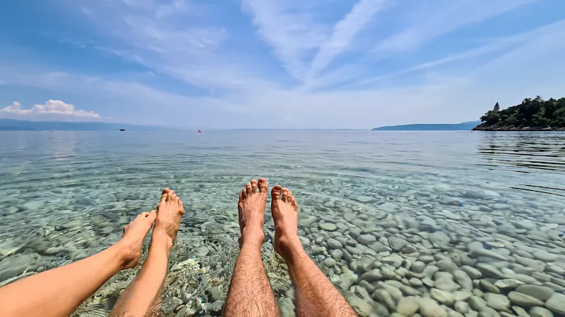 Fire fødder og ben i vandet. Blå himmel og småsten i vandet.