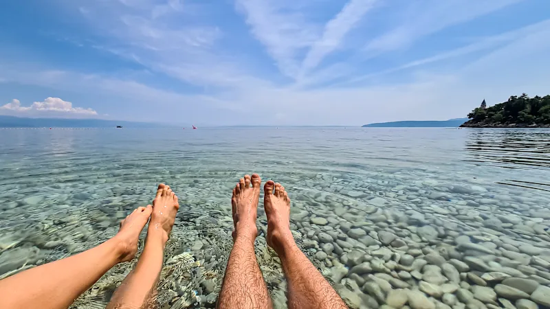 Vier Füße und Beine im Wasser. Blauer Himmel und Kieselsteine im Wasser.