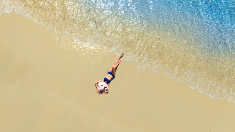Aerial view of beautiful young woman in black swimsuit lying on sandy beach near blue sea with waves at sunset