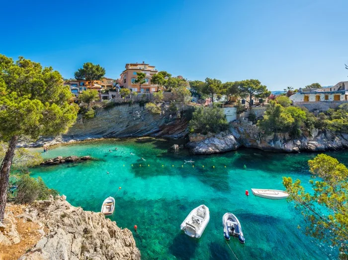 Både flyder i det klare blå vand ved stranden Cala Fornells på Mallorca i Spanien, omgivet af et malerisk kystlandskab.