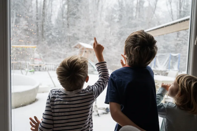 Drei Kinder stehen an einem Fenster und schauen auf die verschneite Landschaft.