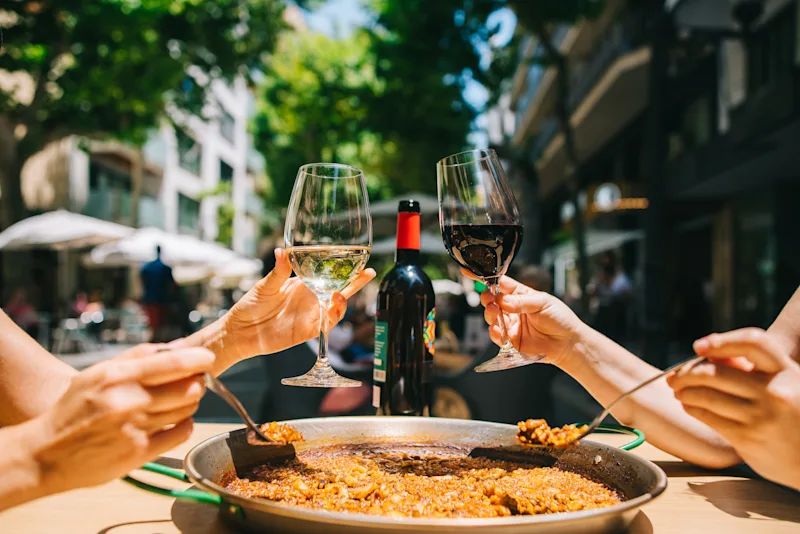 To piger spiser paella og drikker vin på en restaurant, omgivet af en hyggelig atmosfære.