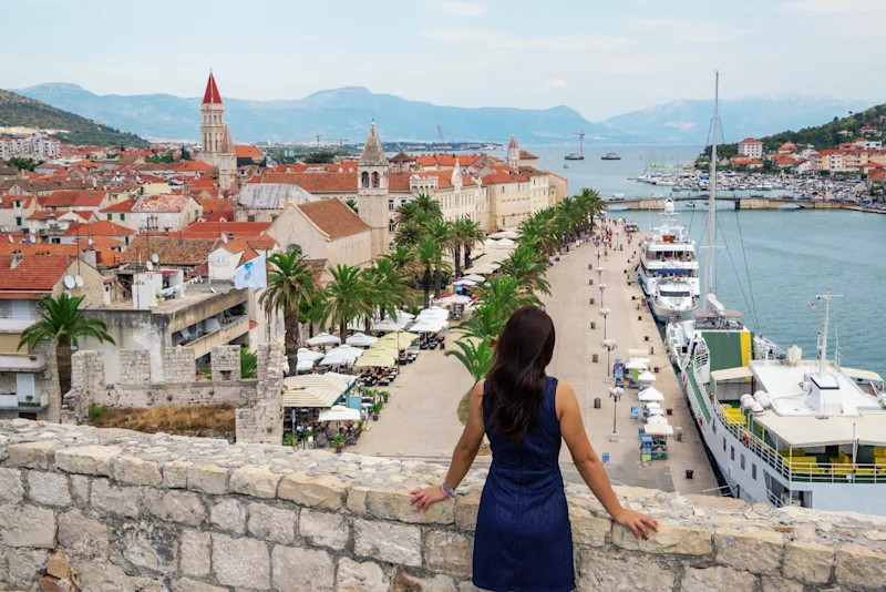 En kvinde kigger ud over promenaden i Trogir med palmer. Der ligger både ved kajen.