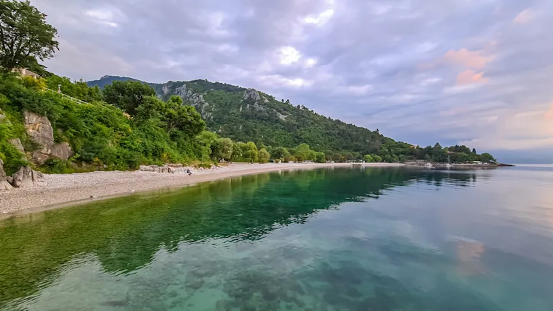 En stenet strand ved kysten af Medveja i Kroatien. Middelhavet er roligt og klart. Ved kysten ligger en frodig skov med en lille by. Himlen er gul.