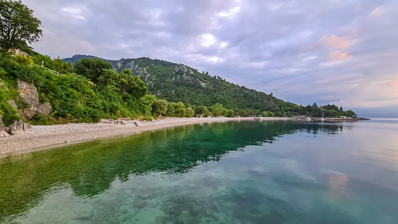 Ein steiniger Strand an der Küste von Medveja in Kroatien. Das Mittelmeer ist ruhig und klar. Am Ufer befindet sich ein üppiger Wald mit einer kleinen Stadt. Der Himmel ist gelb gefärbt.