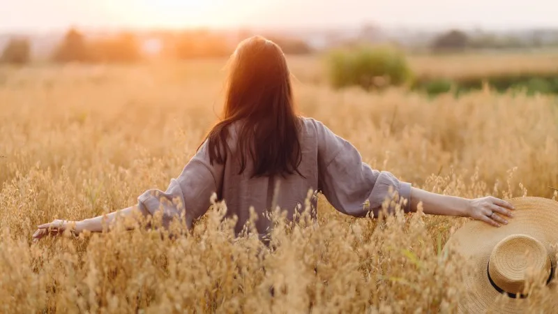 Stylish woman with straw hat standing in oat field in sunset light. 