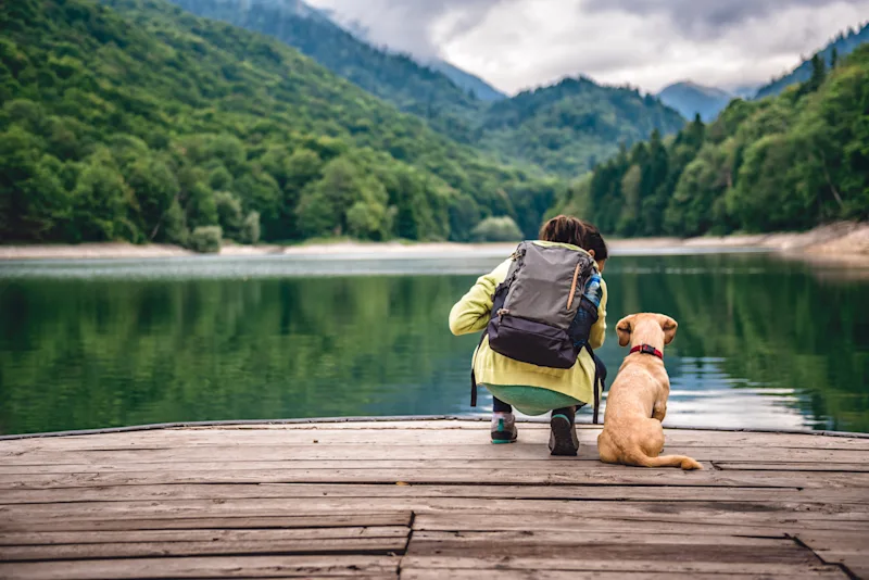 Ein Mann sitzt mit einem Hund auf einem Steg und blickt über das Wasser auf die Berge.