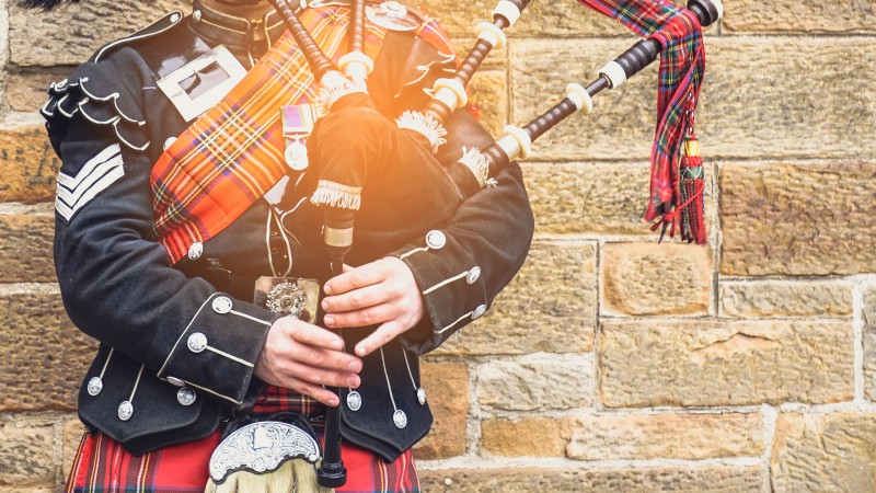 Scottish bagpiper dressed in traditional red and black tartan dress stand before stone wall. 