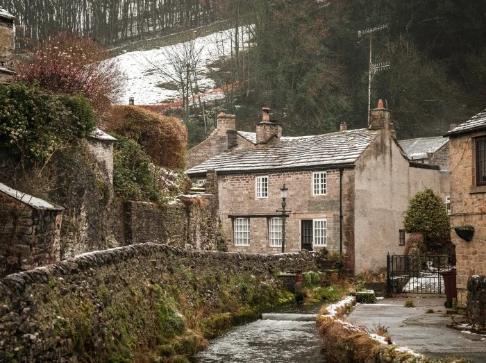 Castleton Hope Valley, Derbyshire Beautiful old rural idyllic village cottages in the Peak District river peacefully flowing during winter with snow on hills ...