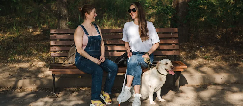 Two friends sitting on a bench together & one of them has a guide dog