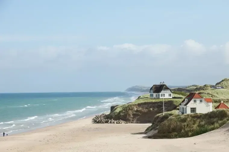 Udsigt over havet fra en strand med to, hvide sommerhuse, som dem du kan leje i Danmark med Novasol.