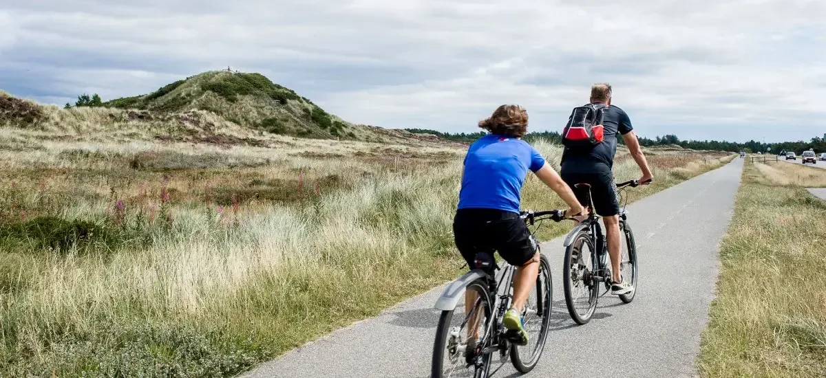 To cyklister på en cykelsti i det grågrønne hedelandskab ved Blåvand, hvor de nyder en aktiv ferie i sommerhus med Novasol.