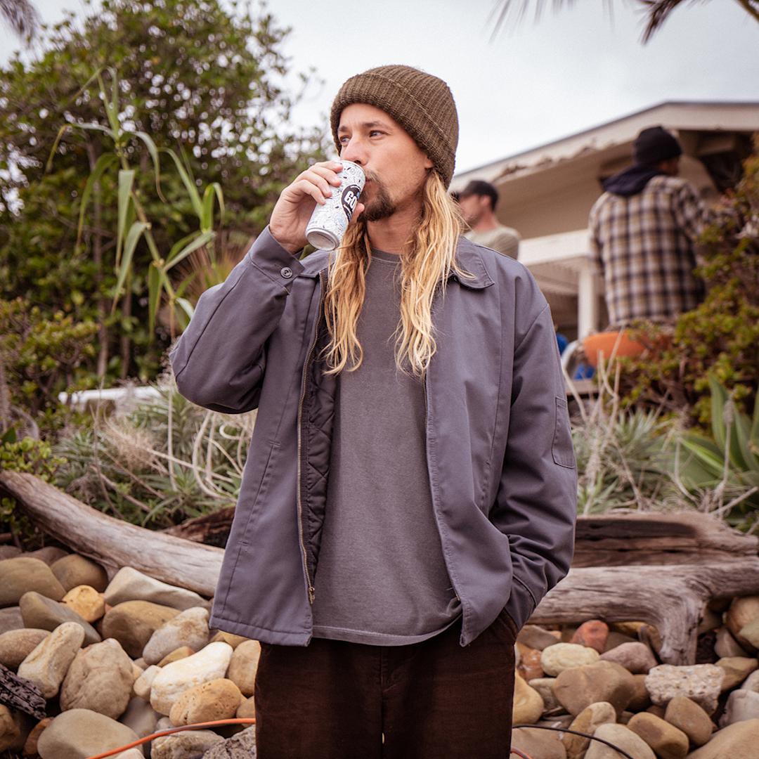 A long haired man in a beanie standing outside drinking a can of Goko