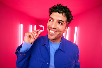 [CRO] Man - Brunette - Curly hair - Smile - Aligner - Looking right - Blue - Jacket - T-shirt - Pink - Light - Background - Mobile
