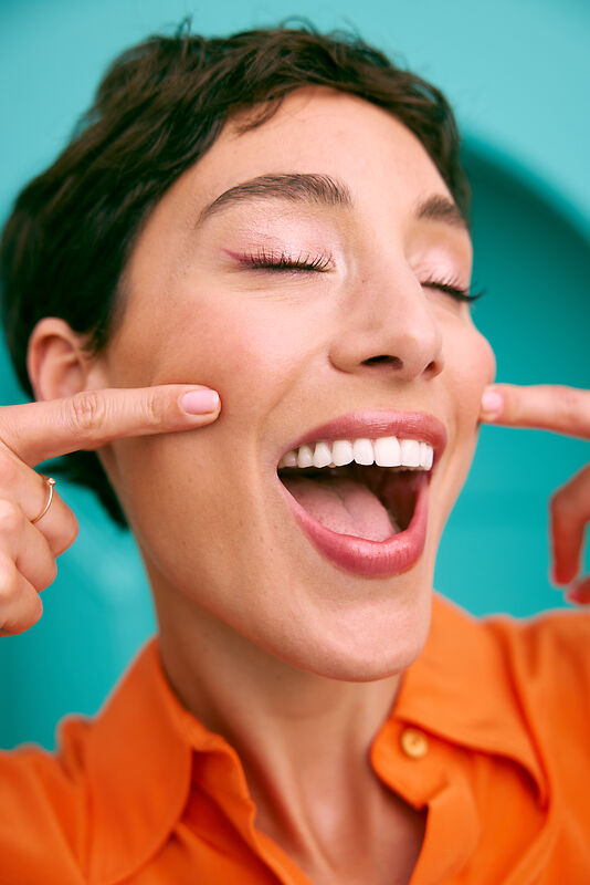 [CRO] Woman - Orange T-shirt - Closed eyes - Mouth Open - Smile - Fingers on the face - Blue - Background - Desktop