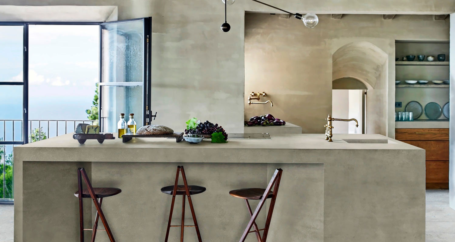 Vaalea betonikeittiösaareke pronssihanoineen, puujakkaroita ja merinäköala avoimesta ikkunasta
