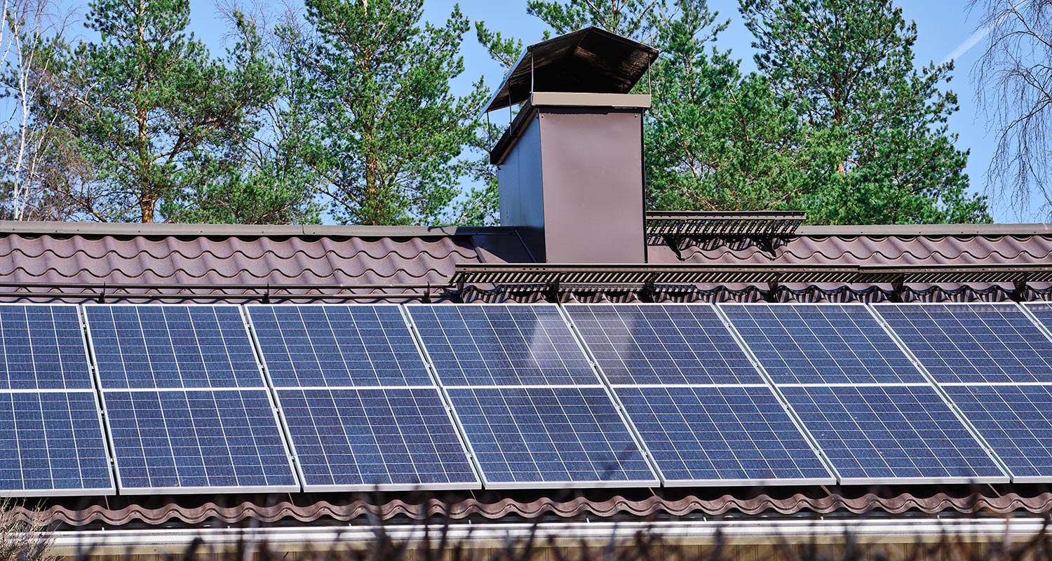 Sinisiä aurinkopaneeleja ruskealla peltikatolla, taustalla havumetsä ja kirkas sinitaivas.