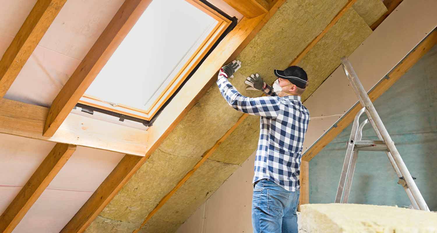 Rakennustyömies asentaa keltaista eristettä puupalkkien väliin vinokaton kattoikkunan alle.