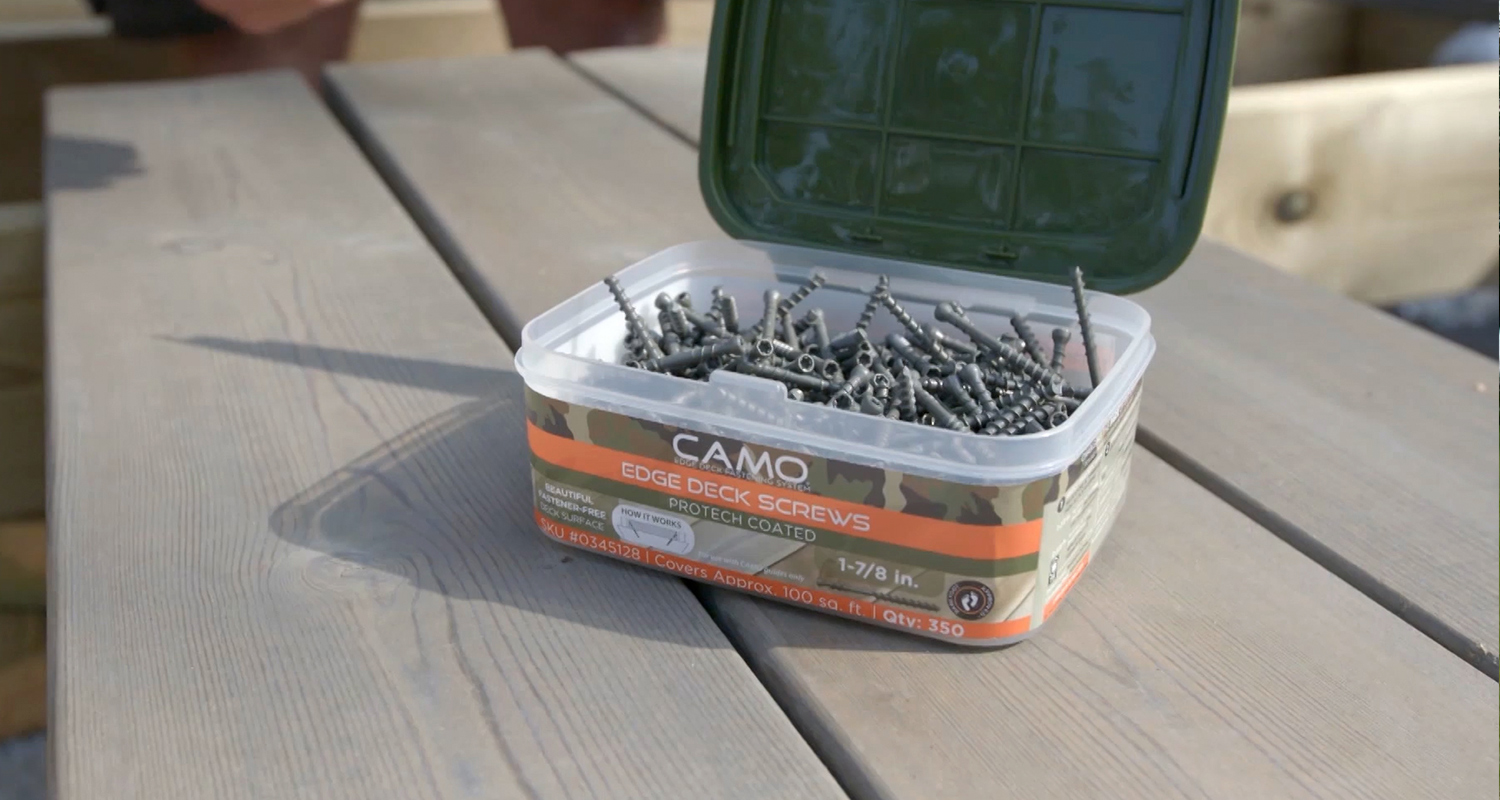 Avoin muovilaatikko oranssi camo-kuvioin täynnä tummia terassiruuveja harmaiden puulautojen päällä