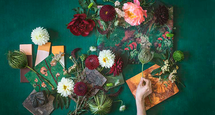 Vihreällä pinnalla käsi asettelee valkoisia, viininpunaisia ja vaaleanpunaisia kukkia