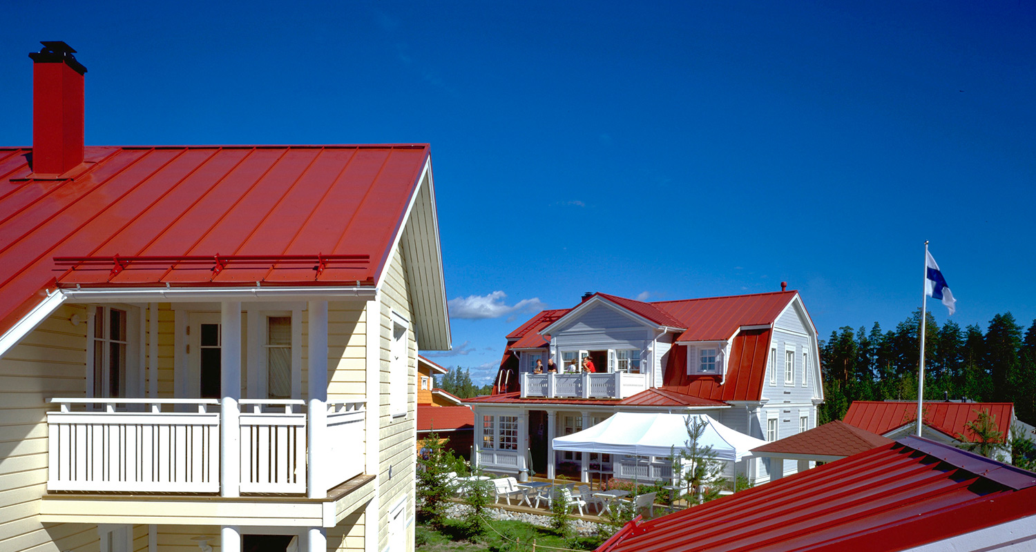 Vaalea talo punaisella katolla, taustalla valkoinen huvila ja liehuva Suomen lippu