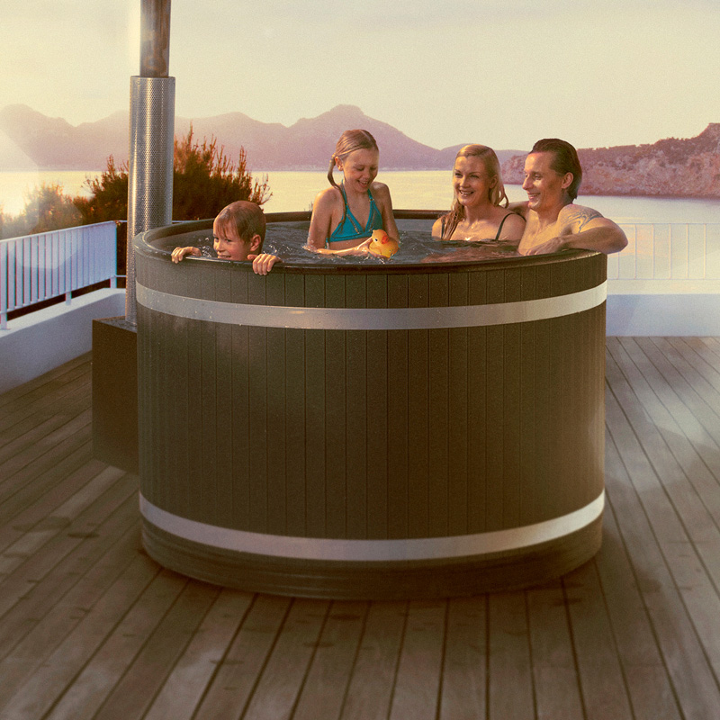 Nelihenkinen perhe porealtaassa puuterassilla järven ja vuorten edessä auringonlaskussa.