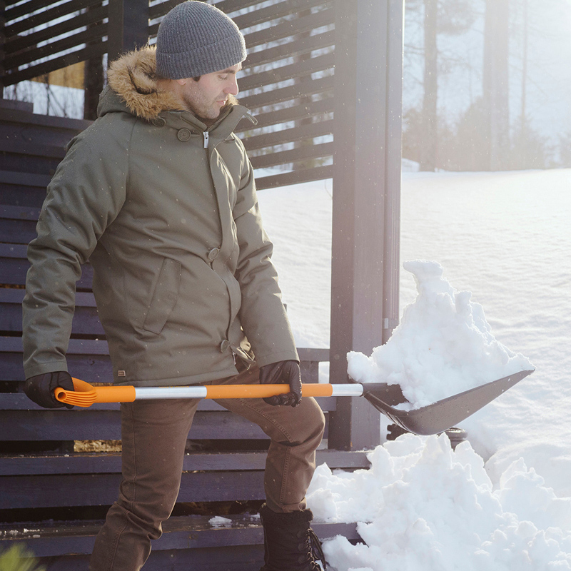 Henkilö vihreässä talvitakissa ja harmaassa pipossa lapioi lunta oranssinvarteisella lapionkauhalla