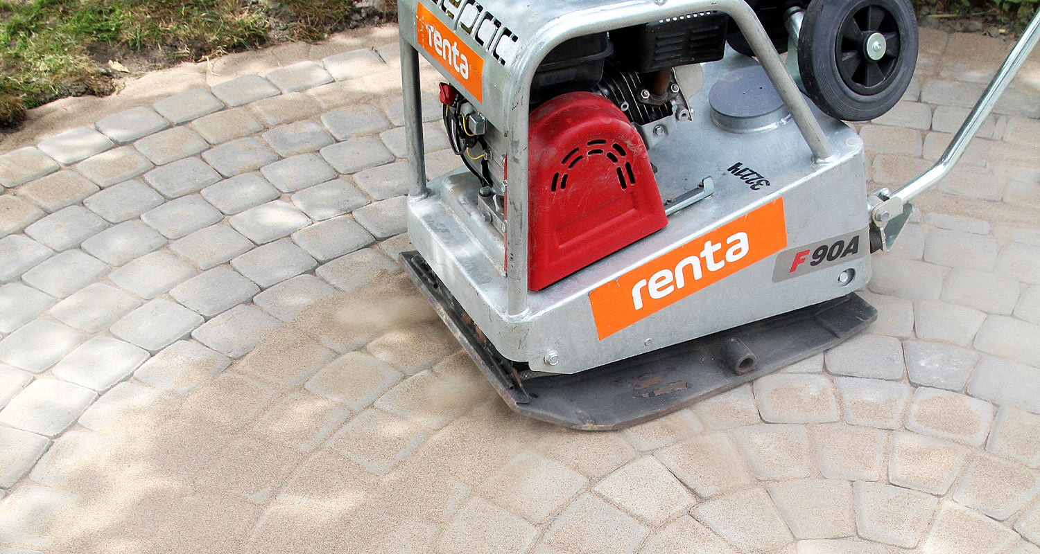 Hopeanharmaa rengasjyrä punaisella kotelolla ja oranssilla logolla tasoittaa soraa pihakivillä.