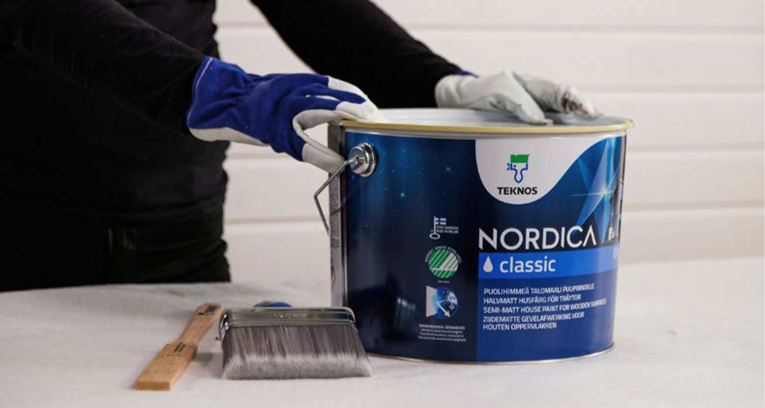 Työhansikkaisiin puetut kädet avaavat sinisen Teknos Nordica maalipurkin, sivellin pöydällä.