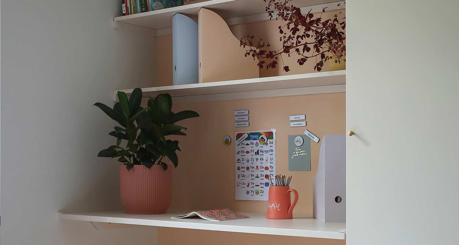 Valkoinen hylly persikkaseinällä, pinkki ruukku ja vihreä kasvi, oranssi kynämuki ja aakkostaulu