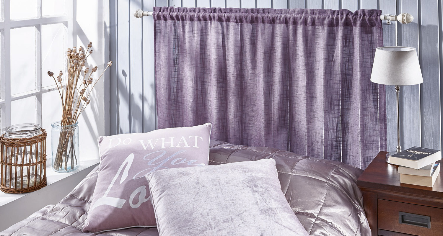 Vaaleanvioletit verhot ja päiväpeitto, kuivatut kukat ikkunalaudalla ja yöpöytä lamppuineen