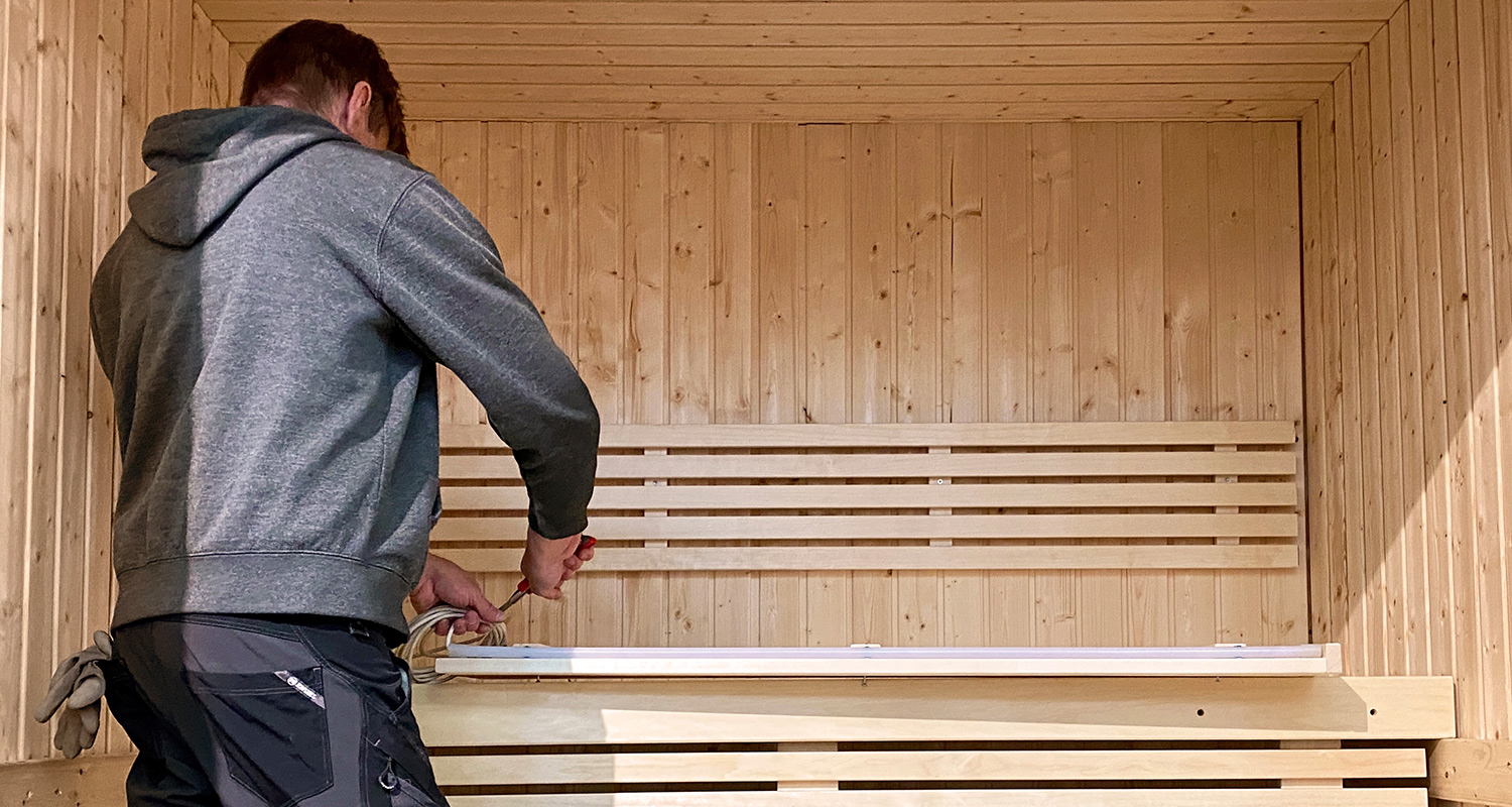 Mies harmaassa hupparissa ruuvaa valolistaa vaaleansävyisessä puusaunassa.