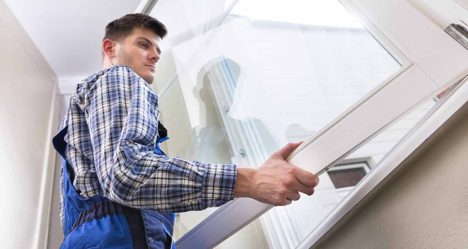 Rakennusmies sinisissä haalarissa asentaa valkoista ikkunankehystä valoisassa sisätilassa