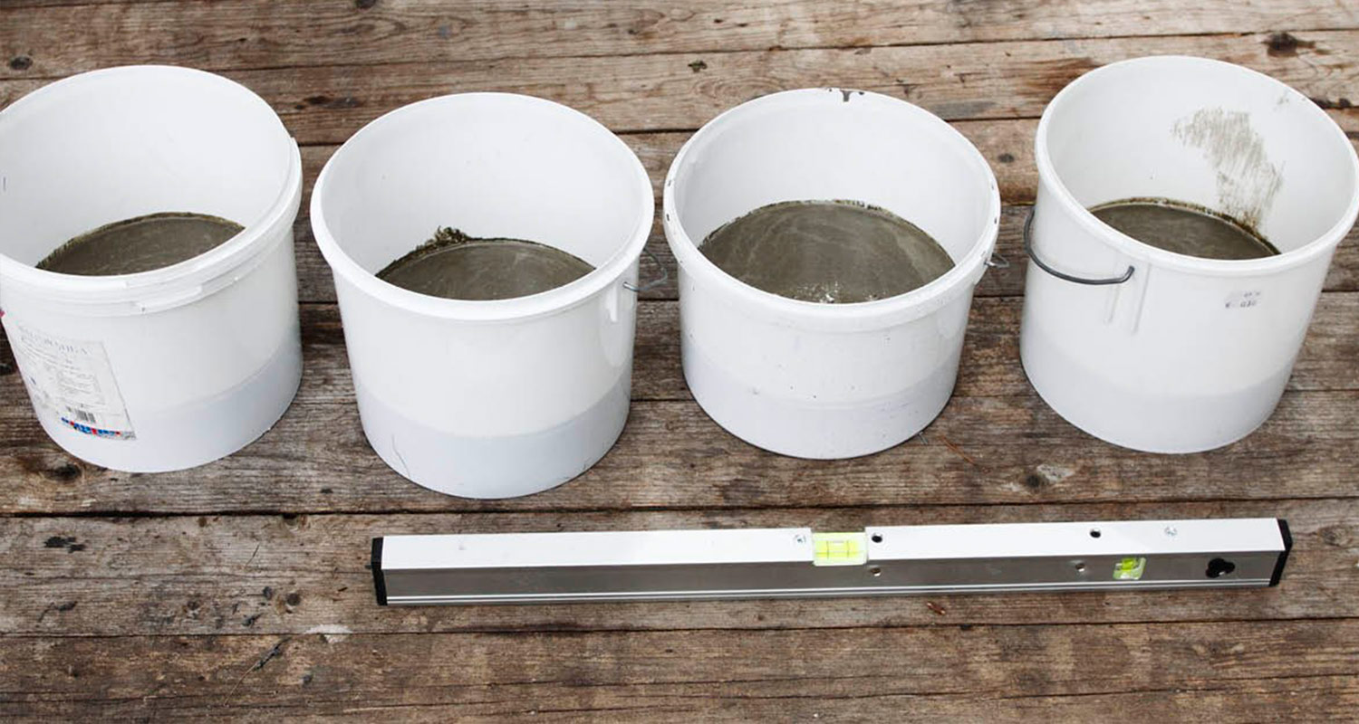 Neljä valkoista ämpäriä harmaata seosta puisella alustalla, etualalla hopeanvärinen vesivaaka.