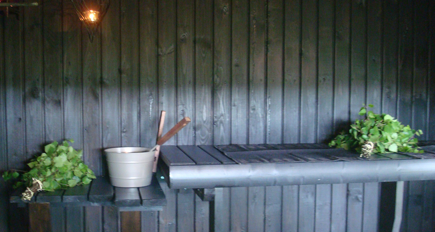 Musta puupaneliseinä, penkillä vihdat ja harmaa kiulu puukauhalla, yläkulmassa lämmin valo