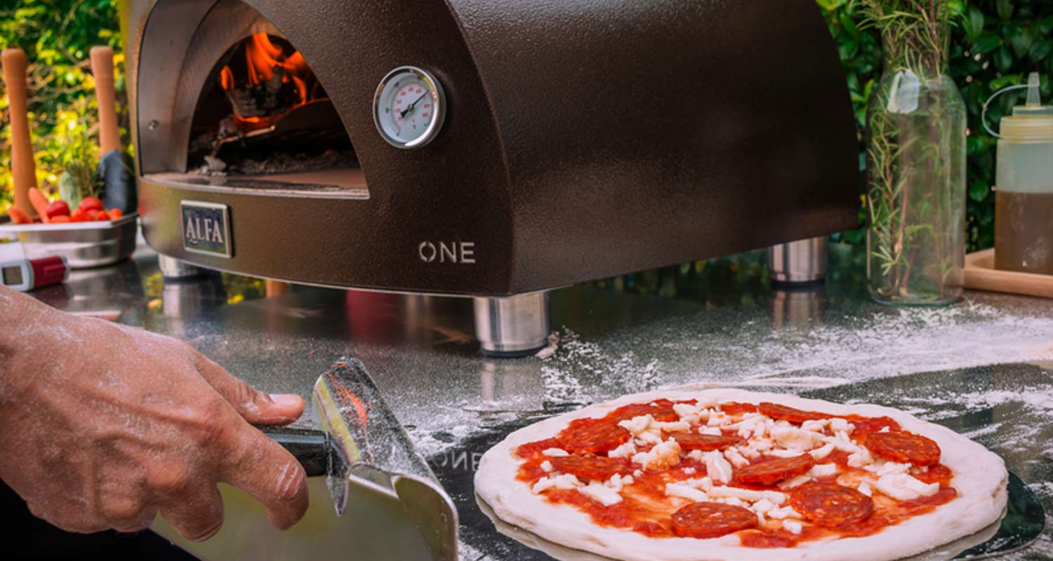 Jauhotettu käsi pizzalapiolla siirtää pepperoni-juustopizzaa pöydällä, taustalla ruskea uuni.