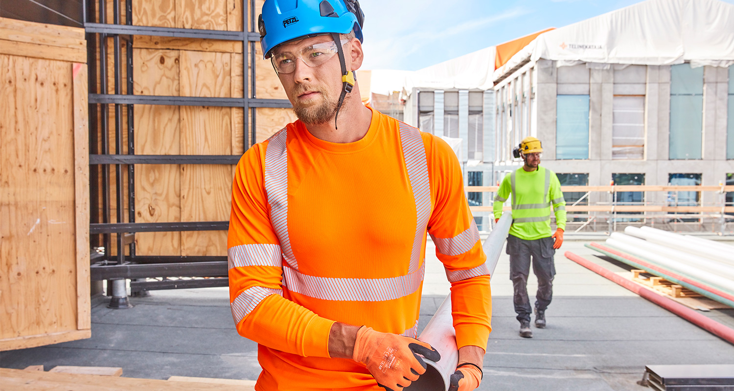 Kaksi rakennusmiestä kantaa raskasta ja pitkää rakennusmateriaalia. Miehillä on yllä K-Raudann PROF Workwear -työvaatteet.