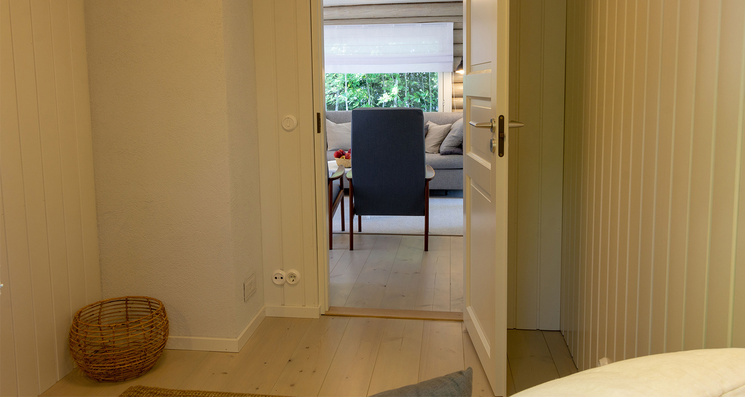 Vaalea puukoti: lattialla kori, avoin ovi, näkymä siniseen nojatuoliin ja harmaaseen sohvaan.