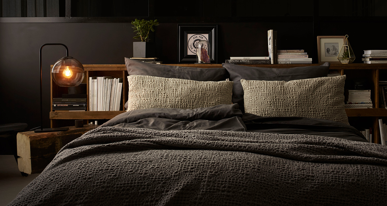 Tummassa makuuhuoneessa harmaa tekstiilipeitto, vaaleat tyynyt, lämmin lasilamppu ja puuhylly