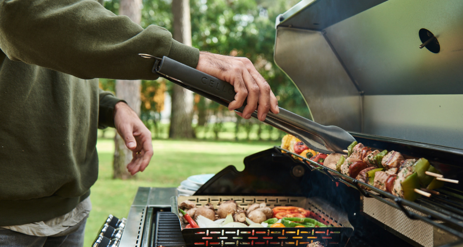 Mies vihreässä paidassa kääntää tangoilla grillissä liha- ja vihannesvartaita puutarhassa.