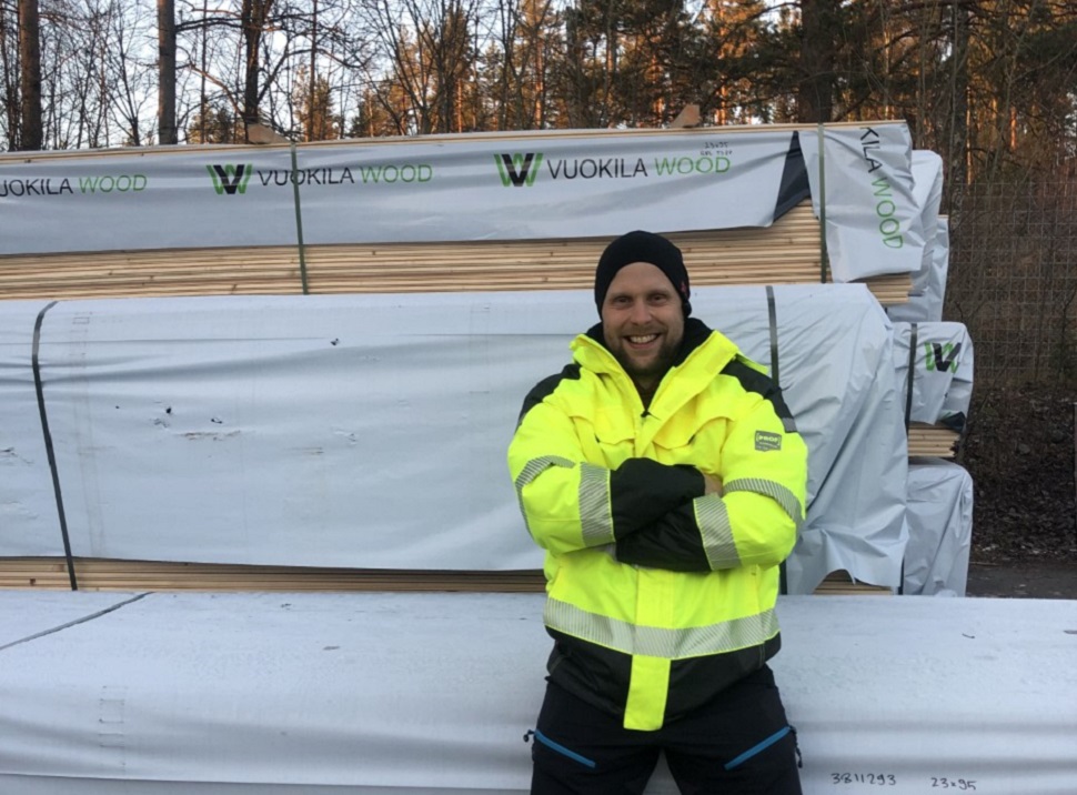 Keltainen heijastintakki ja pinotut puupaketit Vuokila Wood -logoilla metsätaustalla