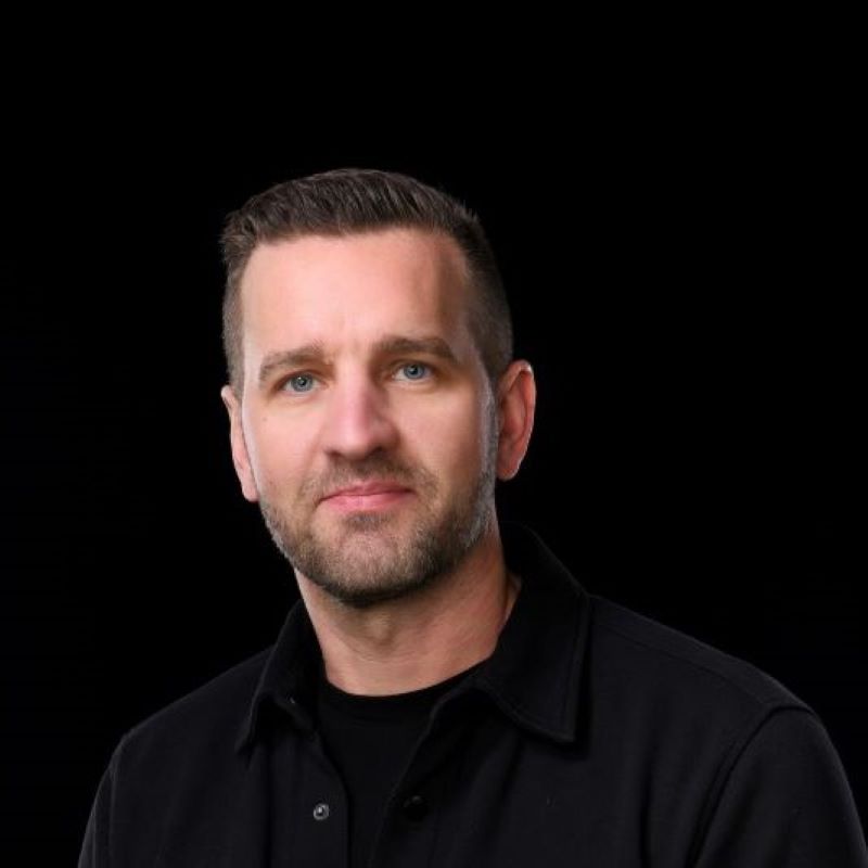 Mies mustassa bleiserissä ja kauluspaidassa yksiväristä mustaa taustaa vasten.